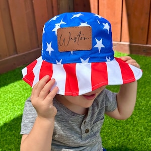 May include: A blue and white star-patterned bucket hat with a red and white striped brim. The hat has a brown leather patch with the name "Weston" on it. The child is also wearing matching red, white, and blue shorts.