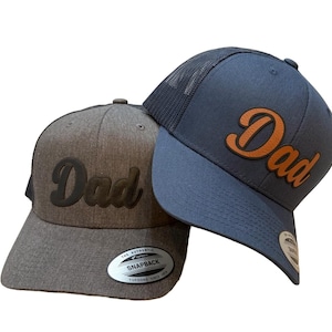 Op de afbeelding: Twee baseballpetten, één grijze en één marineblauwe, elk met het woord "Dad" in een contrasterende kleur. De grijze pet heeft een zwart "Dad"-patch, terwijl de marineblauwe pet een bruine "Dad"-patch heeft. Beide petten hebben een gebogen klep.