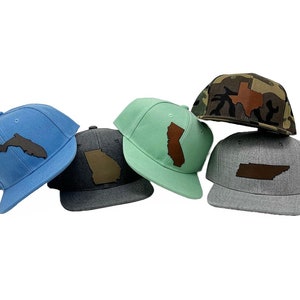 Può includere: Cinque cappellini da baseball con diversi contorni di stati in pelle marrone sul davanti. I cappellini sono di vari colori, tra cui blu, verde, grigio e mimetico.