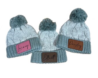 May include: Three blue knit beanies with pom poms and personalized leather patches. The patches are brown, pink, and brown with the names "Paisley", "Lainey", and "Elliott" written in black.