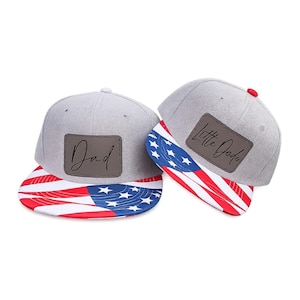 May include: Two grey baseball caps with flat brims featuring a red, white, and blue American flag pattern. Each cap has a rectangular brown leather-like patch on the front. One patch is inscribed with the word Dad and the other with Little Dude.