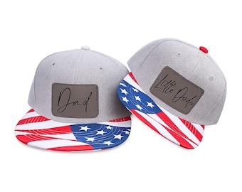 May include: Two grey baseball caps with flat brims featuring a red, white, and blue American flag pattern. Each cap has a rectangular brown leather-like patch on the front. One patch is inscribed with the word Dad and the other with Little Dude.