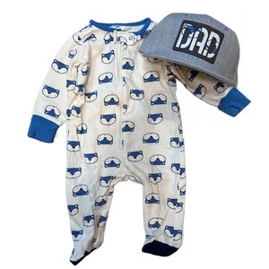 May include: A white baby onesie with a blue fox print and a blue zipper. The onesie has blue trim around the sleeves and legs. A grey baseball cap with a black patch that says "DAD" is included.