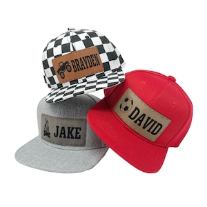Op de afbeelding: Drie gepersonaliseerde baseballpetten met verschillende ontwerpen. De eerste pet is zwart-wit geruit met een bruin patch waarop "BRAYDEN" staat en een monstertruck-afbeelding. De tweede pet is rood met een bruin patch waarop "DAVID" staat en een voetbal-afbeelding. De derde pet is grijs met een bruin patch waarop "JAKE" staat en een kampvuur-afbeelding.