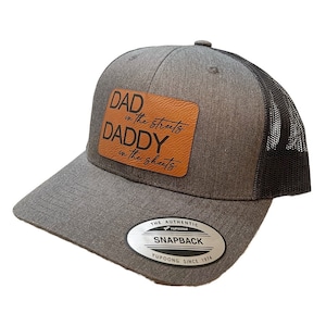 Puede incluir: Una gorra de camionero gris y negra con un parche de cuero marrón que dice "DAD in the streets DADDY in the sheets". La gorra tiene una malla negra en la parte posterior y una etiqueta blanca que dice "THE AUTHENTIC YUPOONG SNAPBACK YUPOONG SINCE 1974".