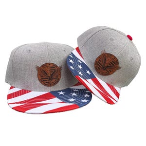 May include: Two grey baseball caps with flat brims featuring a red, white, and blue American flag pattern. Each cap has a circular brown leather patch on the front displaying an eagle graphic and the text 1776, 2026, and 250 Years of Freedom.