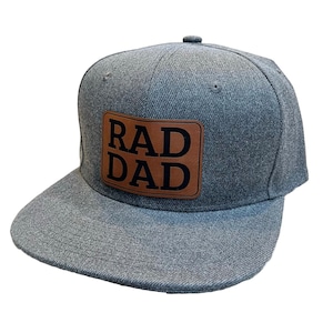 Puede incluir: Una gorra de béisbol gris y marrón con un parche de cuero que dice "RAD DAD" en letras negras.