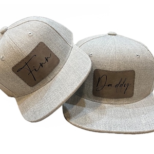 May include: Two gray flat-brimmed baseball caps. One cap has a brown leather patch with the name "Finn" in black script. The other cap has a brown leather patch with the word "Daddy" in black script. Both caps have a structured design.