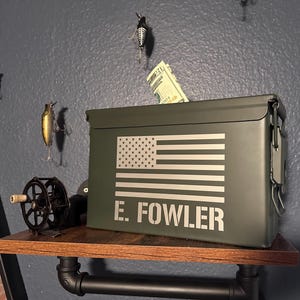 May include: An olive green metal ammo box with a white American flag design and the name "E. Fowler" printed on the front. A stack of U.S. currency is visible inside the box. The box is on a wooden shelf.