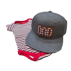 Puede incluir: Un body de bebé a rayas grises y rojas con una gorra de béisbol gris que tiene un parche de cuero marrón con la palabra "DAD" en rayas rojas y blancas.