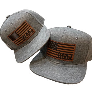 May include: Two gray flat-brimmed baseball caps with brown leather patches. Each patch features a brown American flag design with initials. One reads "WMJ" and the other "SMJ".