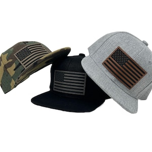 May include: Three flat-brimmed baseball caps with American flag patches. One cap is camouflage, another is black, and the third is gray. Each cap has a rectangular patch with the flag design.