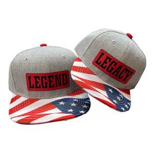 May include: Two gray baseball caps with American flag designs. One cap says "LEGEND" and the other says "LEGACY".