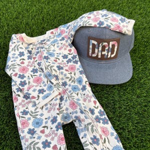 Op de afbeelding: Een baby-slaappakje met bloemenprint, ritssluiting en een denim baseballpet met het woord "DAD" op een leren patch. Het slaappakje heeft een crèmekleurige achtergrond met roze en blauwe bloemen.