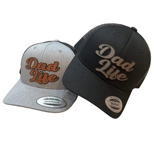 Op de afbeelding: Twee trucker caps met de tekst "Dad Life" in een retro lettertype. De ene is grijs met oranje tekst en de andere is zwart met zilveren tekst.