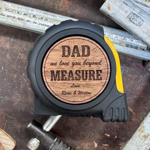 May include: A black and yellow tape measure featuring a circular wooden inlay on the side. The wood is engraved with the text: DAD we love you beyond MEASURE Love. Remi & Weston. The tool has a black rubberized casing and a yellow accent.