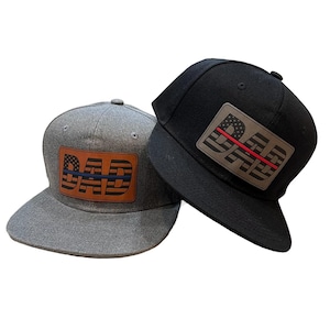 Op de afbeelding: Twee baseballpetten met een leren patch waarop "DAD" staat met een Amerikaans vlagontwerp en een dunne rode of blauwe lijn over de vlag. De ene pet is grijs en de andere is zwart.