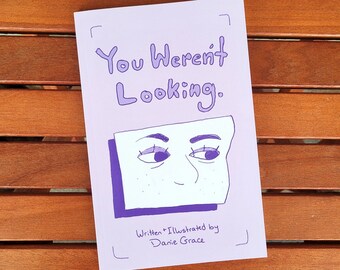 Könnte beinhalten: Ein lila Buch mit dem Titel "You Weren't Looking". Das Cover zeigt eine Illustration von Augen und einer Nase in einem weißen Rechteck. Der Text "Written + Illustrated by Danie Grace" steht darunter. Das Buch liegt auf einer Holzoberfläche.