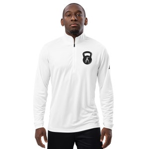 May include: White long-sleeve athletic shirt with a black zipper and a black kettlebell graphic on the left chest. The kettlebell graphic features a silhouette of a person lifting the kettlebell.