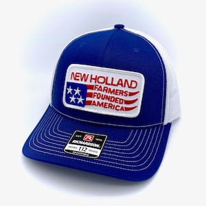 May include: A royal blue trucker hat with a white mesh back. The front of the hat features a white patch with a red, white, and blue American flag design and the text "NEW HOLLAND FARMERS FOUNDED AMERICA". The hat also has a black and red label with the text "RICHARDSON 112 ORIGINAL TRUCKER ADJUSTABLE".