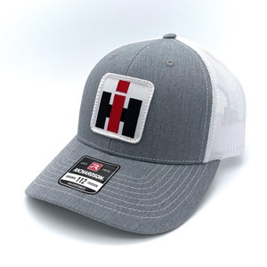 May include: Gray and white trucker hat with a patch featuring a red, black, and white logo. The hat has a curved brim and a mesh back. The tag reads "RICHARDSON ORIGINAL 112 TRUCKER ADJUSTABLE".