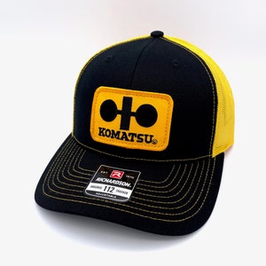 May include: Black and yellow trucker hat with a Komatsu logo patch. The patch is yellow with a black Komatsu logo and a black circle with a white line.