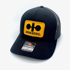 May include: Black trucker hat with a yellow and black Komatsu logo patch. The hat is adjustable and has a mesh back.