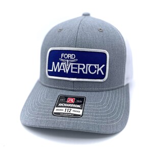 May include: A gray and white trucker hat with a blue rectangular patch featuring the word "FORD" above "MAVERICK" in white lettering. The hat has a curved brim and a mesh back. The Richardson brand label is visible.