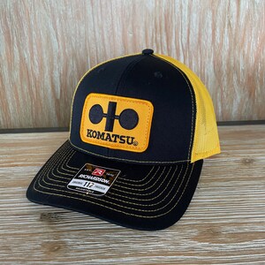 May include: Black and yellow trucker hat with a yellow patch that says "KOMATSU" and a logo of two circles connected by a line. The hat has a black brim and a yellow mesh back.