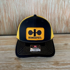May include: Black and yellow trucker hat with a Komatsu logo patch. The hat is adjustable and has a black mesh back.