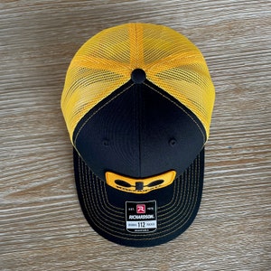 May include: Black and yellow trucker hat with a yellow logo and the text "KOLPAK" and "EST. 1970 RICHARDSON. ORIGINAL TRUCKER 112 ADJUSTABLE".