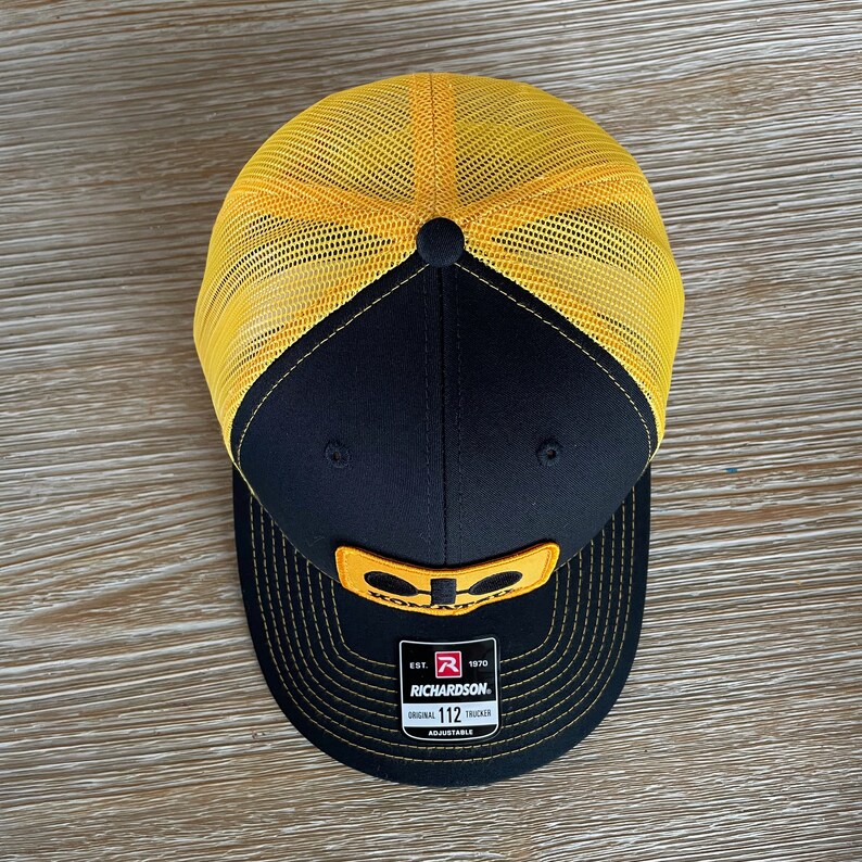 May include: Black and yellow trucker hat with a yellow logo and the text "KOLPAK" and "EST. 1970 RICHARDSON. ORIGINAL TRUCKER 112 ADJUSTABLE".