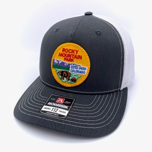 May include: A gray and white trucker hat with a yellow patch featuring a bear and the text "Rocky Mountain Park Estes Park Colorado". The hat has a black and red label with the text "Richardson 112 Original Trucker Adjustable".
