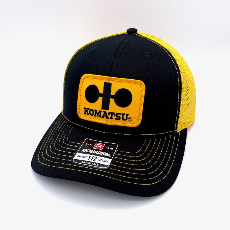 May include: Black and yellow trucker hat with a Komatsu logo patch. The patch is yellow with a black Komatsu logo and a black circle with a white line.