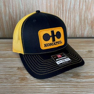 May include: Black and yellow trucker hat with a Komatsu logo patch. The hat is adjustable and has a mesh back.