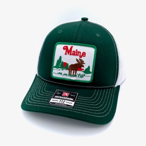 May include: A green and white trucker hat with a patch that says "Maine" and features a moose in a forest scene. The hat is adjustable and has a Richardson 112 label.