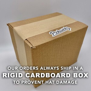 May include: A brown cardboard box with a white label that says "Pickwick's" with an arrow. The box is taped shut. The text below the box reads "Our orders always ship in a rigid cardboard box to prevent hat damage."