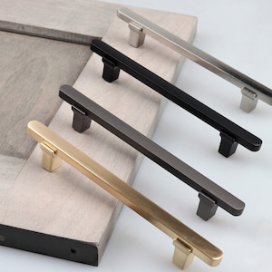 May include: Four cabinet pulls in different finishes: gold, nickel, black, and gunmetal. The pulls are all the same size and shape, with a rectangular base and a slightly curved handle.