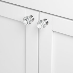 May include: Two clear crystal cabinet knobs with a faceted design and silver metal bases are mounted on white cabinet doors.