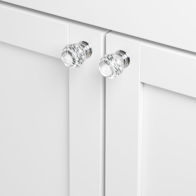 May include: Two clear crystal cabinet knobs with a faceted design and silver metal bases are mounted on white cabinet doors.