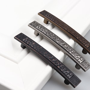 May include: Three cabinet pulls in different finishes: black, silver, and bronze. Each pull has a hammered texture and a square design at each end.
