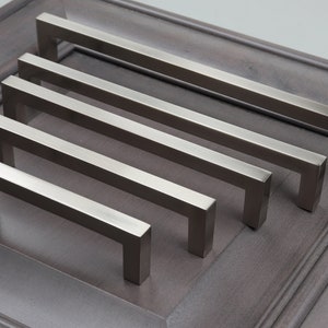 May include: Five brushed nickel cabinet pulls of varying lengths, arranged in a row on a dark gray surface.