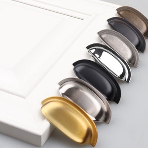 May include: A set of six cabinet pulls in various finishes, including brass, nickel, chrome, and black. The pulls are all the same shape, a curved, slightly rounded rectangle.