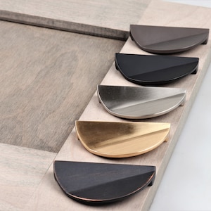May include: Five half-circle cabinet pulls in different finishes: black, brushed nickel, gold, oil-rubbed bronze, and dark gray. The pulls are arranged in a row on a light wood surface.