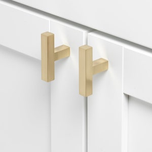 May include: Two gold cabinet pulls with a rectangular shape and a T-shaped design. The pulls are mounted on white cabinet doors.