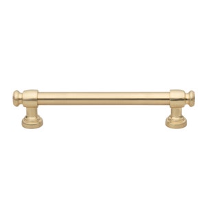 May include: A gold-colored cabinet pull with a cylindrical bar and decorative end pieces. The pull is approximately 5 inches long and features a classic design, suitable for drawers and cabinets.
