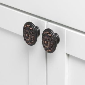 May include: Two black and copper cabinet knobs with a decorative floral design. The knobs are mounted on white cabinet doors.