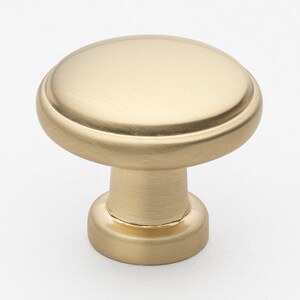 May include: A brushed gold cabinet knob with a round top and a slightly raised edge. The knob has a simple, elegant design.