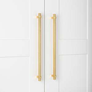 May include: Two gold cabinet pulls with a square design at the top and bottom. The pulls are mounted on a white door.