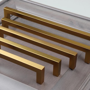 May include: A collection of five gold-colored, square-shaped cabinet pulls of varying lengths. The pulls are arranged in a row, showcasing their different sizes. The hardware is set against a gray background, highlighting the metallic finish.